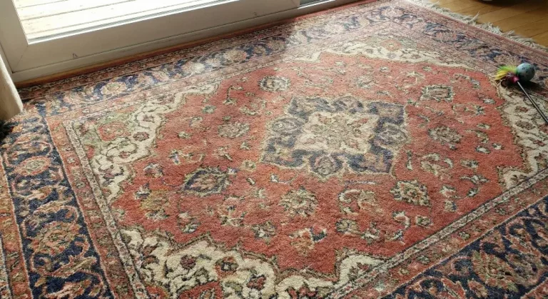 Photo of a vintage carpet in the eastern style with a red-blue color, in the summer The wool is placed on the wooden top of the window, with the cat toy in the corner.
